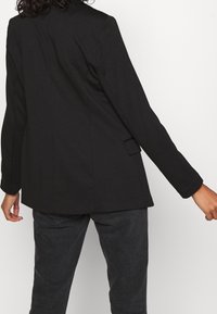 Musta blazer, jossa on muotolaskettu malli, yksittäinen halkio takana, pitkät hihat ja hienovaraisesti teksturoitu kangas. Yhdistetty tummanharmaisiin housuihin.