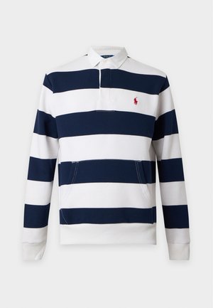 Jeune homme portant une chemise à manches longues rayée noir et blanc, un pantalon cargo beige et des baskets blanches, debout les mains dans les poches.