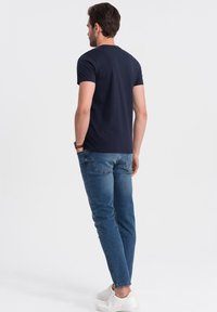 Námořnické modré tričko s krátkým rukávem v kombinaci se světle modrými džíny. Model má na sobě bílé tenisky, stojí s rukama v kapsách, pohled z této strany.