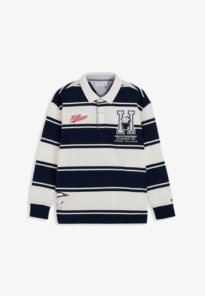 Uśmiechnięty chłopiec ubrany w granatowo-białą, paskowaną, długorękawową koszulkę polo z logo Snoopy i Tommy Hilfiger, stojący na białym tle.