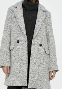 Vrouw draagt een getextureerde lichtgrijze wollen jas over een donkergrijze geribbelde coltrui, staand tegen een effen witte achtergrond.