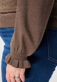 Bruine gebreide trui met een glinsterende textuur en een gerimpelde manchetkant. Gecombineerd met donkerblauwe denim.