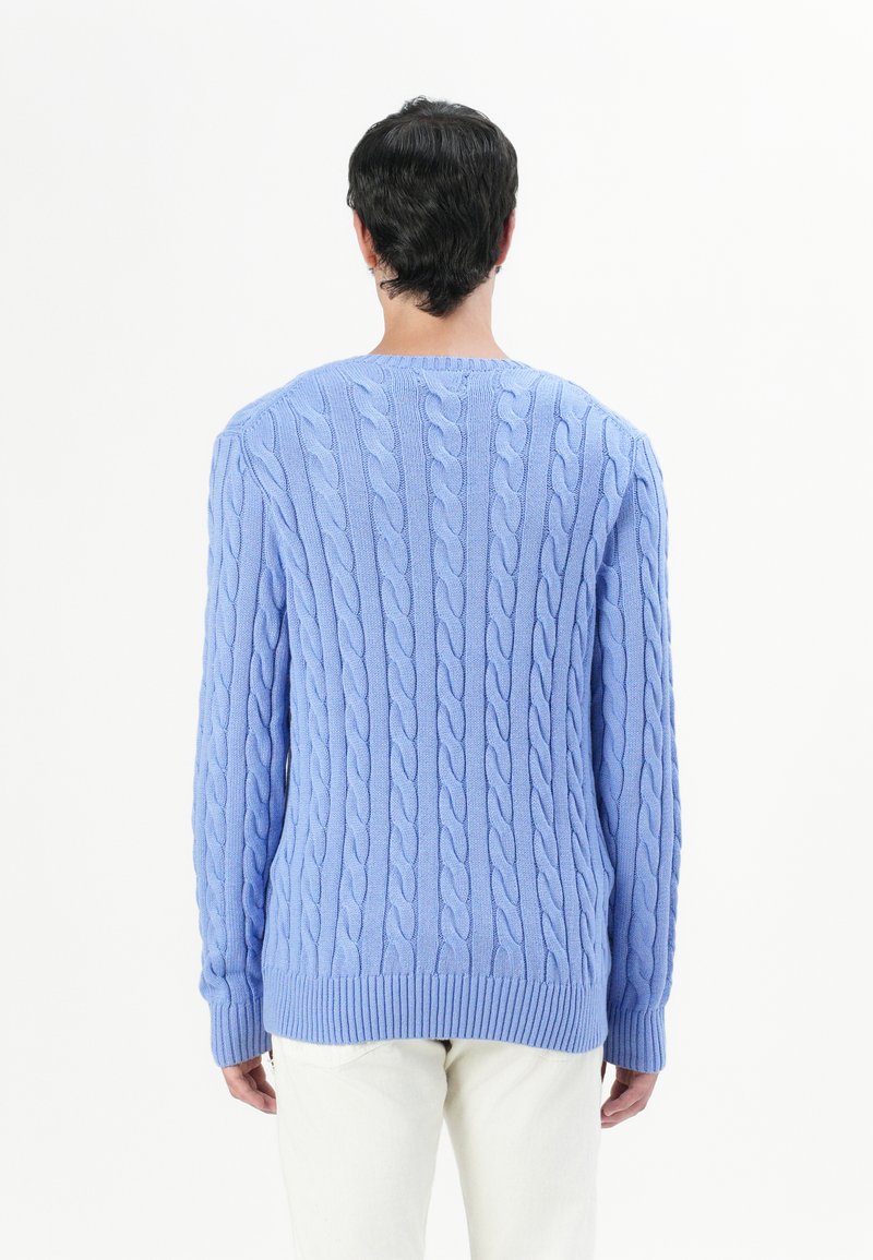 Maglione a maglia blu cielo con motivo a treccia testurizzato, scollo a girocollo e polsini e fondo a costine, indossato sopra pantaloni di colore chiaro.