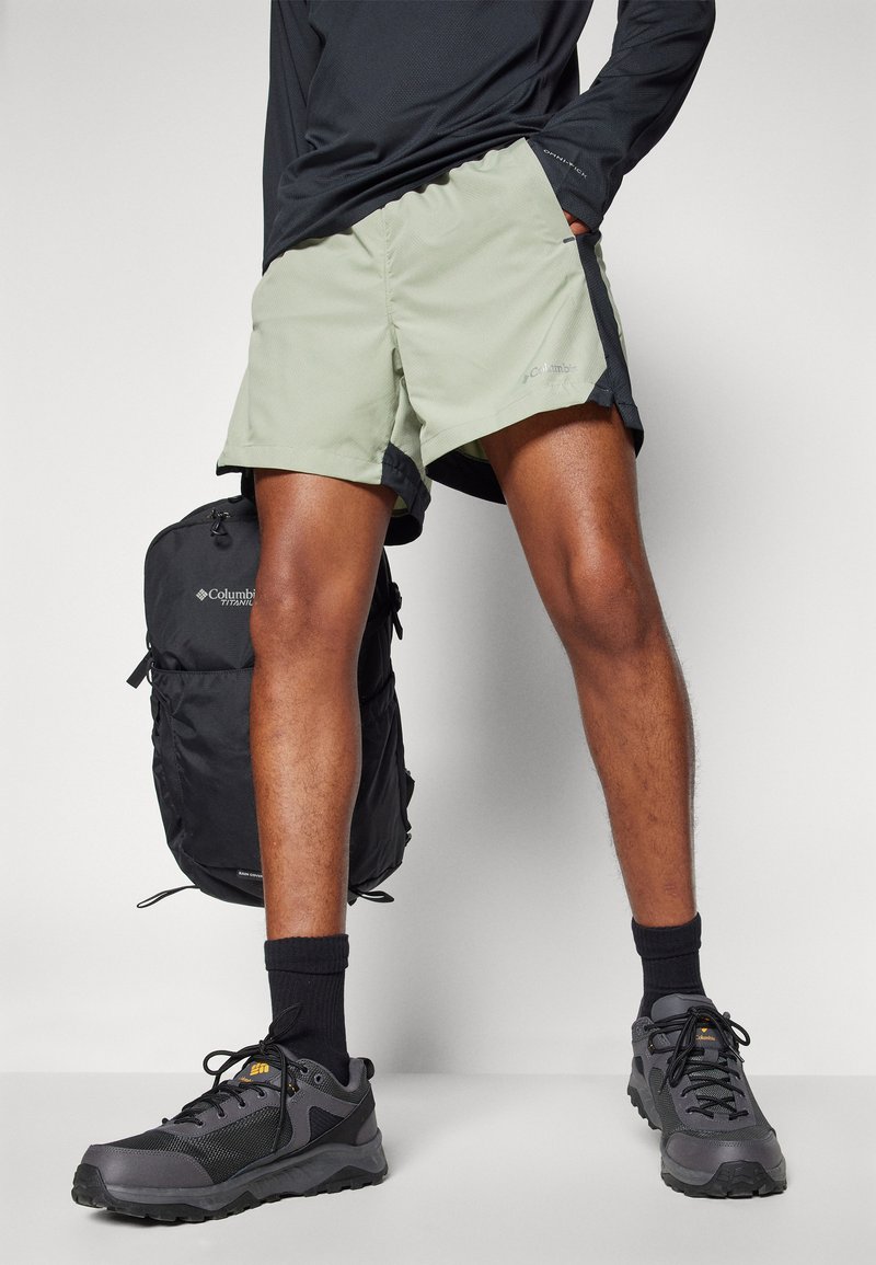 Pantalones cortos verde claro con ribete negro, combinados con zapatos de senderismo grises y calcetines negros hasta el tobillo. Una mochila negra es visible detrás.