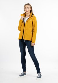Gul softshell jakke med høj krave, to sidelommer og lynlås. Sammen med mørkeblå tætsiddende jeans og grå sneakers.