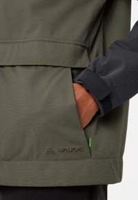 Grüne wasserfeste Jacke aus strukturiertem Material, mit elastischen Bündchen, einer Seitentasche und einem gestickten "Vaude"-Logo.