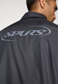 Sort jakke med en glat tekstur, der har et fremtrædende gråt "SPURS" logo på ryggen og et klassisk kravedesign.
