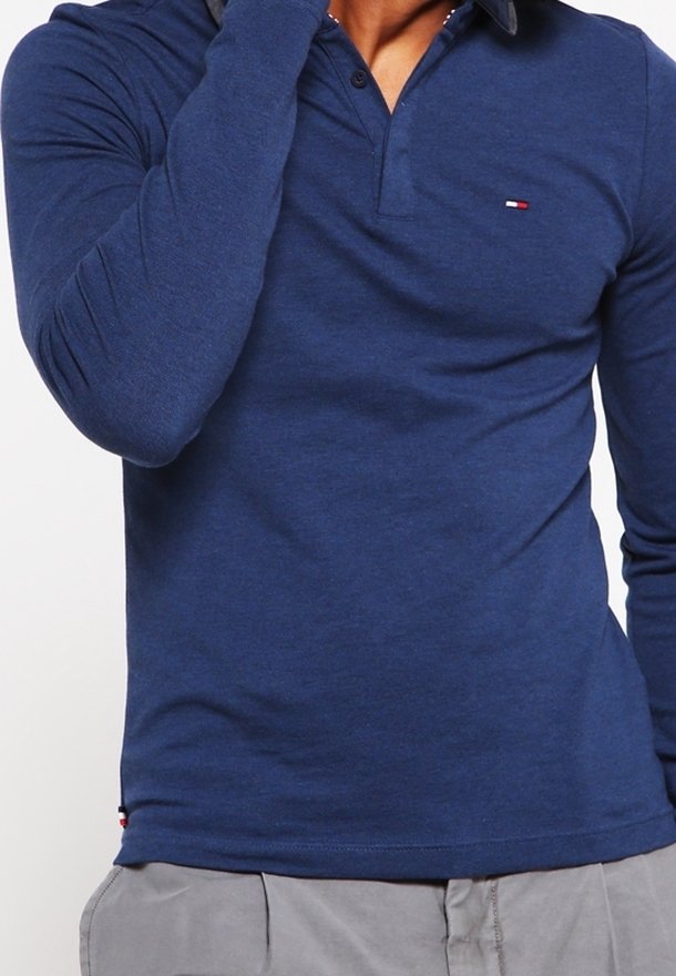 Homme portant un polo bleu ajusté à manches longues avec un petit logo sur la poitrine et un pantalon gris clair, main près du visage.