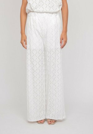 Femme portant un pantalon blanc à jambes larges en tissu ajouré à motifs losanges et des sandales beiges ouvertes, debout devant un fond uni.