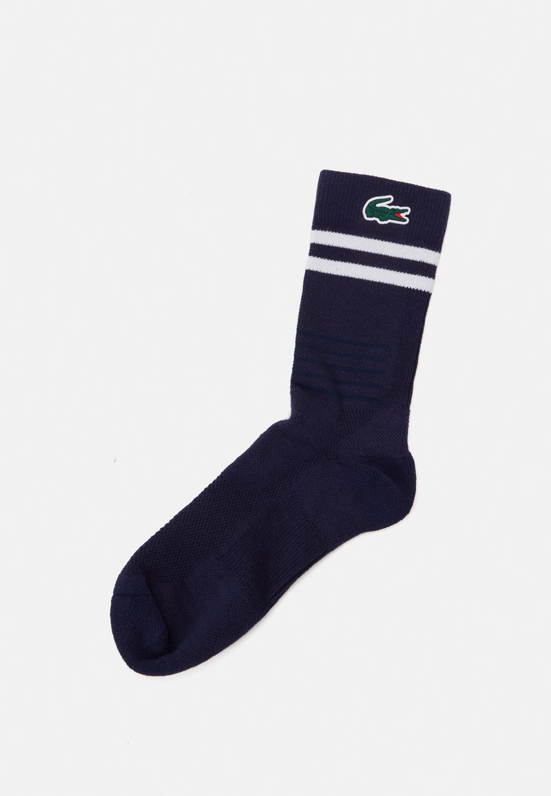 Temno modre bombažne nogavice z belimi črtami na vrhu. Imajo teksturiran vzorec in izvezen zelen logotip Lacoste na strani.