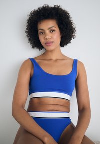 Blå sport-BH og matchende høyhøyde bikini i elastisk stoff, med en hvit bånd med "HUGO"-logo og svart aksentlinje.