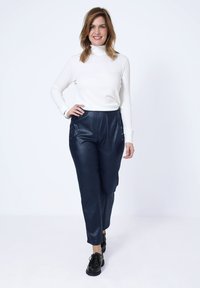 Witte coltrui en marineblauwe neplederen broek met knoopdetails aan de zijkant, gedragen met zwarte schoenen op een neutrale achtergrond.