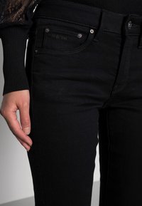 Jeans de mezclilla negra con corte ajustado, que cuentan con bolsillos delanteros, detalles en hardware plateado y una tela suave, con una ligera textura.
