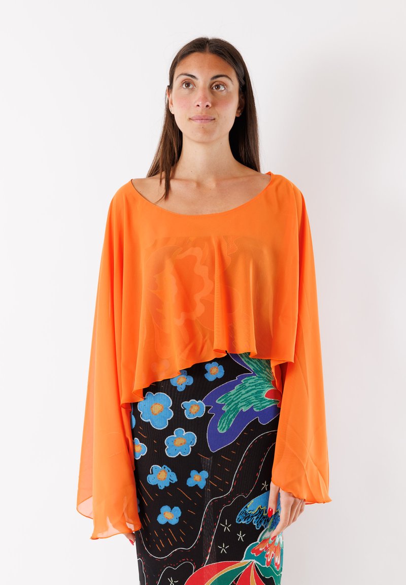 Blusa arancione trasparente con scollatura ampia e maniche fluide, indossata sopra un vestito nero con motivi floreali colorati.