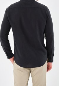 Camisa negra de mangas largas con dobladillo curvo y puños abotonados. Fabricada con tejido texturizado, ajuste entallado, costuras visibles y detalle de yugo en la espalda.