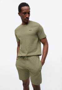 Άνδρας που φοράει ελαιοπράσινο υφασμάτινο κοντομάνικο πουκάμισο και αντίστοιχα σορτς με το λογότυπο «BOSS», στέκεται με τα χέρια στις τσέπες, κοιτάζοντας προς τα κάτω.