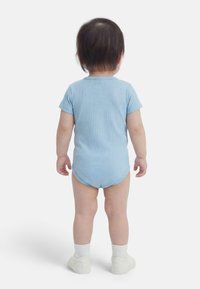 Väikelaps seisab seljaga, seljas lühikeste varrukatega sinine soonikkoes bodi, jalas valged sokid ja valged kingad, taust on ühevärviline valge.