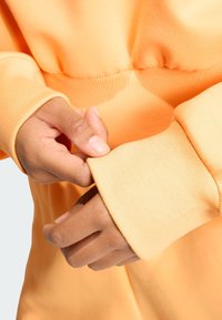 Pomarańczowy sweatshirt z prążkowanymi mankietami; dłonie dostosowujące rękaw. Tkanina ma gładką teksturę, podkreślając jej bliskie dopasowanie.