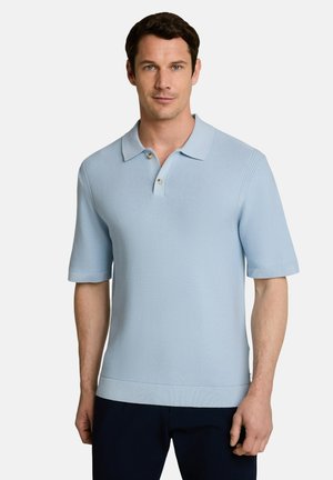 Man with short brown hair wears light blue short-sleeve polo shirt and dark pants, standing against plain light background.