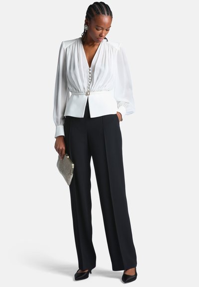 Femme portant un chemisier blanc à manches longues avec des boutons, un pantalon noir à jambes larges, des talons noirs, tenant une petite pochette métallique.