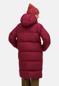 Bordeaux pufferjas met een capuchon, met gevoerde secties. Het ontwerp is lang en iets oversized, gecombineerd met een rode gebreide muts.