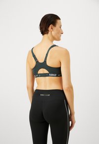 Sostén deportivo de color verde oscuro con tirantes ajustables, un diseño de espalda descubierta y una banda de marca negra. Combinado con leggins negros que presentan un logo.
