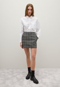 Weißes Hemd mit Knöpfen und einer Tasche, kombiniert mit einem karierten Minirock in Grau und Schwarz. Das Modell trägt schwarze Loafer und knielange Socken.