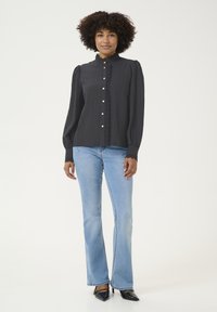 Schwarze Bluse mit hohem Kragen und Rüschendetails, ausgestattet mit Knopfverschlüssen und langen Puffärmeln. Kombiniert mit hellblauen Schlagjeans.