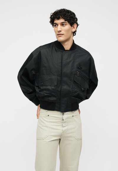 Νέος άνδρας με σγουρά μαλλιά φορώντας μαύρο bomber μπουφάν με πολλές τσέπες και ανοιχτό μπεζ παντελόνι, στέκεται μπροστά σε απλό φόντο.