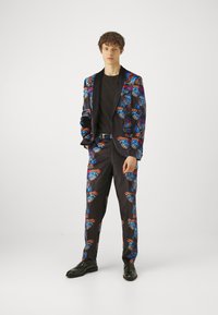 Svart dress med livlig sommerfugltrykk i blått, rødt og oransje; inkluderer en blazer og matchende bukser, kombinert med en svart t-skjorte.