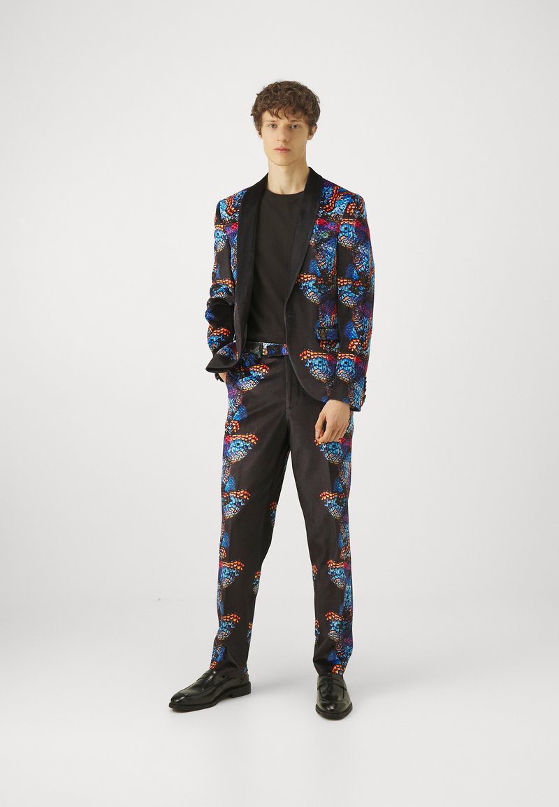 Svart dress med livlig sommerfugltrykk i blått, rødt og oransje; inkluderer en blazer og matchende bukser, kombinert med en svart t-skjorte.