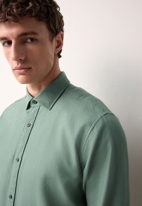 Camicia verde texturizzata con colletto, dotata di chiusura frontale con bottoni e dettagli di cuciture sottili. Tessuto morbido e vestibilità comoda.