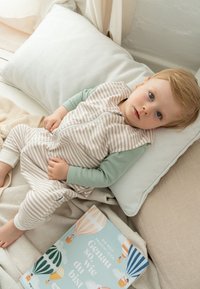 Hellbeige und gestreiftes Babyanzug mit grünen Ärmeln, getragen von einem Kind, das auf neutral gefärbten Kissen sitzt, neben einem illustrierten Buch auf einer weichen Oberfläche.