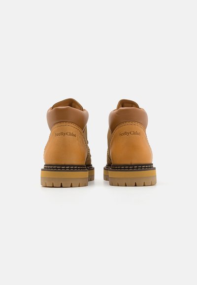 Bottines en cuir marron avec une finition lisse, accents beiges contrastants, talons arrondis, et semelles en caoutchouc crantées. Logo embossé sur le côté.