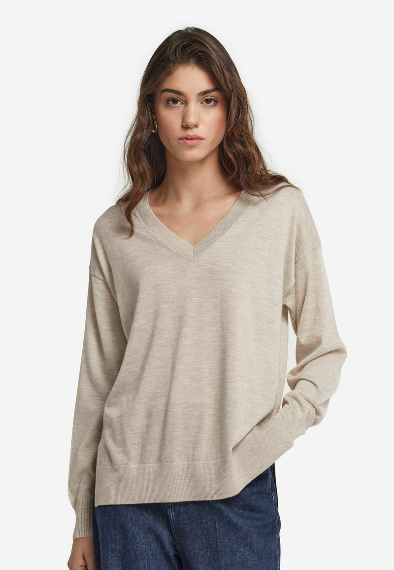 Beżowy, oversize sweter z dekoltem w serek, wykonany z miękkiego, dzianinowego materiału, z długimi rękawami i ściągaczem na dole. Zestawiony z ciemnoniebieskimi spodniami dżinsowymi.