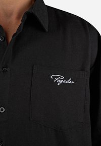 Camisa preta de botões, feita de um tecido texturizado, apresentando um bolso no peito com bordado branco "Pegador". Colarinho clássico e fecho por botões.