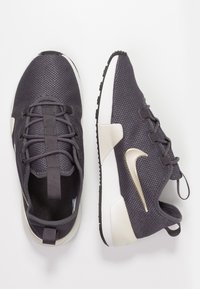 Mörkgrå nätlöparskor med vit sula och guld Nike-logotyp. Har platta snören och flexibel gummisula för grepp.