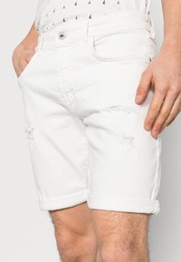 Shorts de mezclilla blanca con dobles dobladillos, con acentos desgastados y bolsillos delanteros. Diseño clásico con cierre de botón.