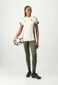 Camisa adidas verde claro com detalhes em verde escuro, combinada com leggings verdes escuras. Modelo segura uma bola de futebol preta e branca.