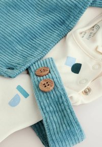 Blaues Cordstoffkleidungsstück mit gerippter Textur, ausgestattet mit Holzknöpfen. Über einem cremefarbenen Einteiler mit bunten geometrischen Mustern drapiert.