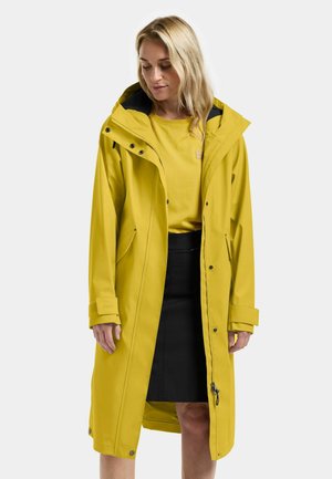 Żółta wodoodporna kurtka przeciwdeszczowa z kapturem, przestronnym krojem, zapięciami na guziki i zamek oraz czarną podszewką. W połączeniu z żółtą bluzką i czarną spódnicą.
