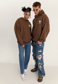 Divi jaunieši, ģērbti saskaņotās brūnās kapučjakās un zilās džinsās, stāv pret vienkāršu fonu; vienam ir saulesbrilles un plēstas bikses.