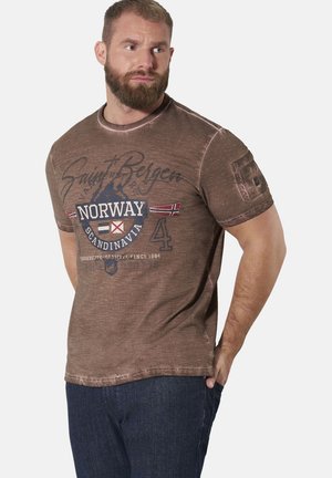 Man with beard wearing brown Norway graphic t-shirt and dark jeans, looking to his left with one hand in pocket against plain background.