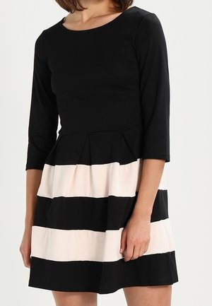 Woman wearing a black dress with three-quarter sleeves and alternating wide black and beige horizontal stripes on the skirt.