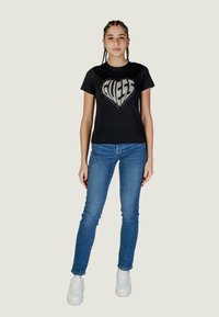 T-shirt in cotone nero con lettering "GUESS" in strass argentati a forma di cuore, abbinato a jeans skinny blu e sneakers bianche.