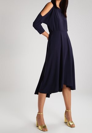 Frau trägt ein marineblaues Midi-Kleid mit transparenten Schultern und goldene Sandalen mit Blockabsatz, steht mit einer Hand in der Hüfte.