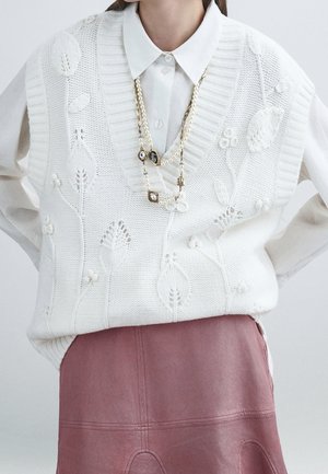 Pull sans manches blanc tricoté avec motifs floraux sur une chemise blanche boutonnée, associé à une jupe en cuir mauve et un collier de perles.
