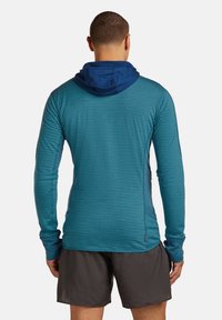 Uomo che indossa un hoodie a maniche lunghe di color teal con cappuccio blu scuro e pantaloni corti grigi scuri, in piedi di spalle su uno sfondo bianco semplice.