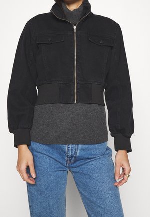 Vrouw die een zwart cropped jas met rits draagt over een grijze trui met blauwe jeans, staand tegen een effen witte achtergrond.