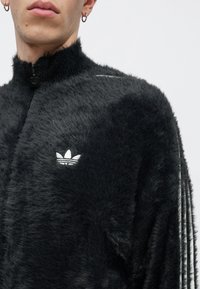 Fekete műszőrme dzseki magas gallérral, cipzáros záródással és fehér Adidas logóval. Csíkos díszítésekkel az oldalán. Puha tapintású.